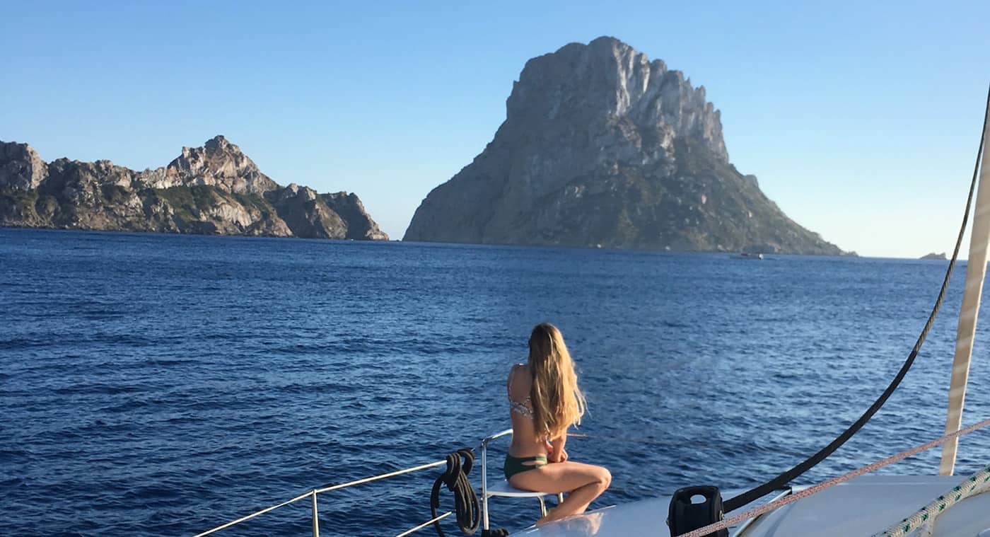 Chica sentada en el balcon de proa del catamaran admirando la isla de Es Vedra