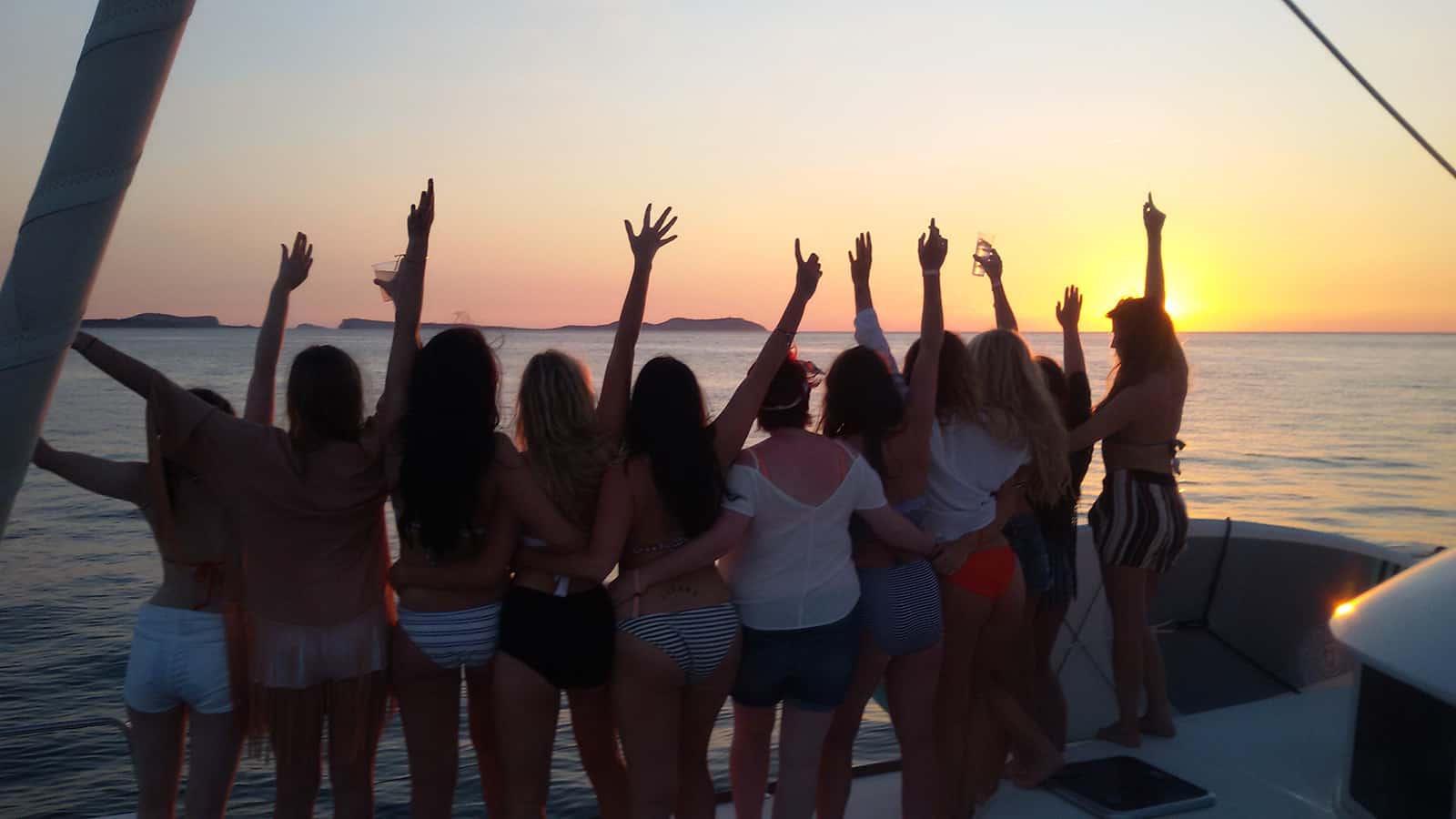 Enterrement de vie de jeune fille sur un catamaran à Ibiza, filles sur le dos levant les bras au coucher du soleil.