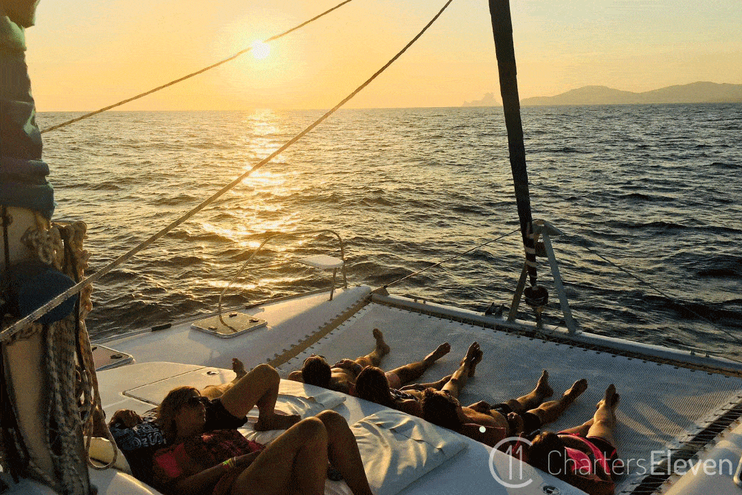 Alquiler catamaran Ibiza día, amigos tumbados en la red mientras se navega con la puesta de sol.