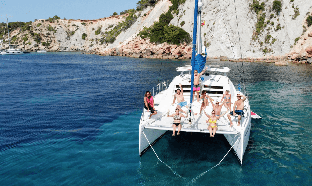 Grupo de amigos en la proa del catamaran de alquiler en Ibiza saludando a cámara.