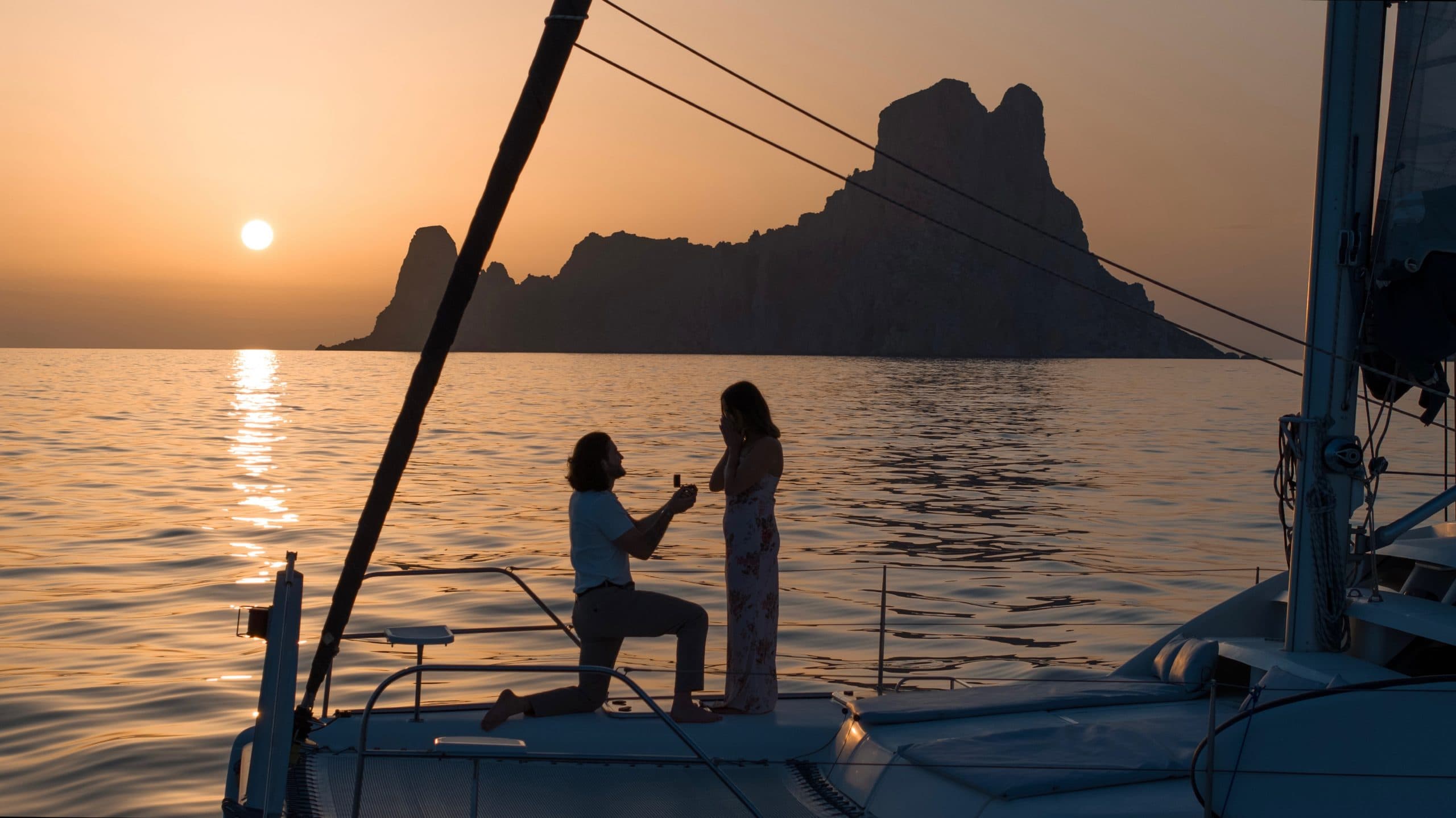 Boy kneeling at the bow of the catamaran when asking for his partner's hand with the sunset and Es Vedra.